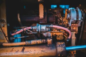 Technician inspecting a noisy hydraulic pump, hoses, reservoir, and pressure lines on industrial equipment.
